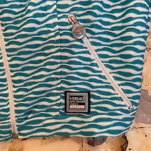 Op de afbeelding: Een blauw-wit gestreept jack met een zilveren rits en een zwart label met de tekst "VERSACE JEANS COUTURE PIECE M. 388520".