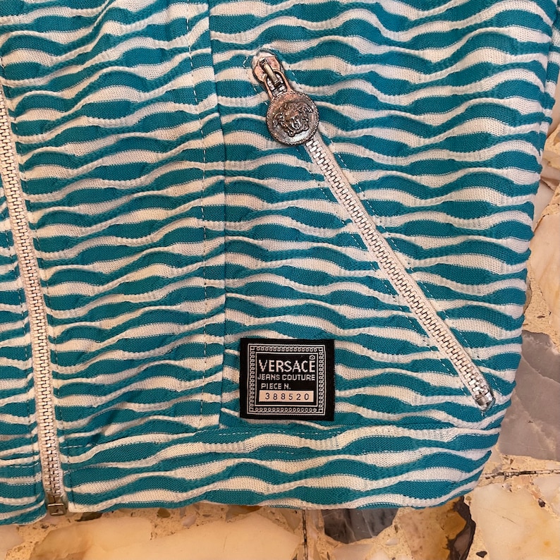 Op de afbeelding: Een blauw-wit gestreept jack met een zilveren rits en een zwart label met de tekst "VERSACE JEANS COUTURE PIECE M. 388520".