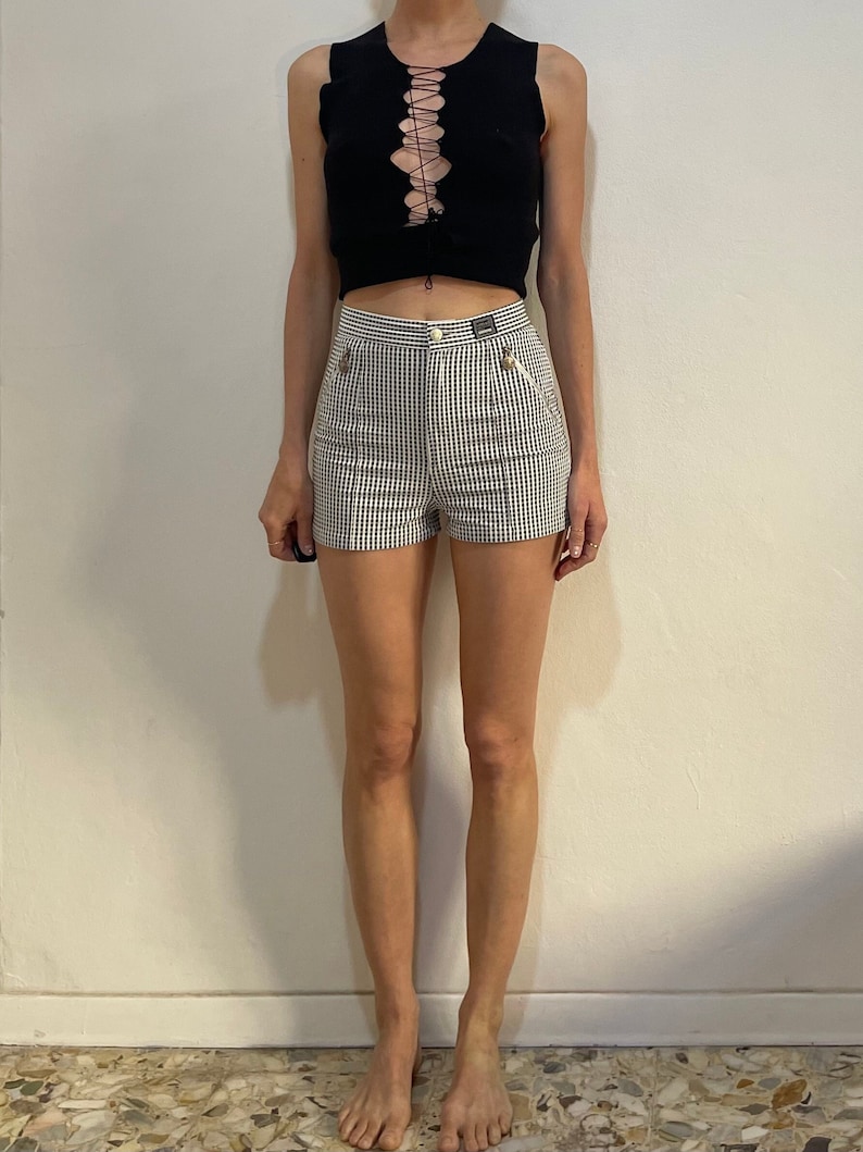 May include: A black sleeveless top with a lace-up front is paired with high-waisted shorts. The shorts feature a black and white checkered pattern, a button closure, and small pockets. The outfit is a casual, yet stylish ensemble.