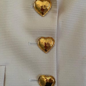 Puede incluir: Primer plano de una chaqueta color crema con tres botones dorados en forma de coraz&oacute;n. Los botones texturizados reflejan la luz, agregando elegancia a la prenda. La chaqueta tiene una sutil textura acanalada.