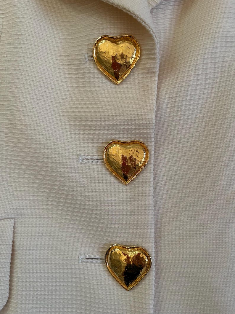 Puede incluir: Primer plano de una chaqueta color crema con tres botones dorados en forma de coraz&oacute;n. Los botones texturizados reflejan la luz, agregando elegancia a la prenda. La chaqueta tiene una sutil textura acanalada.