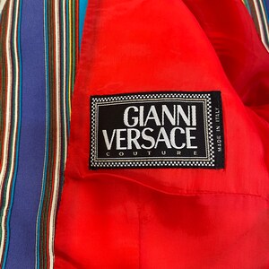 May include: A red fabric with a black label that reads "Gianni Versace Couture" and "Made in Italy". The fabric has a striped pattern in blue, red, yellow, green, and white.