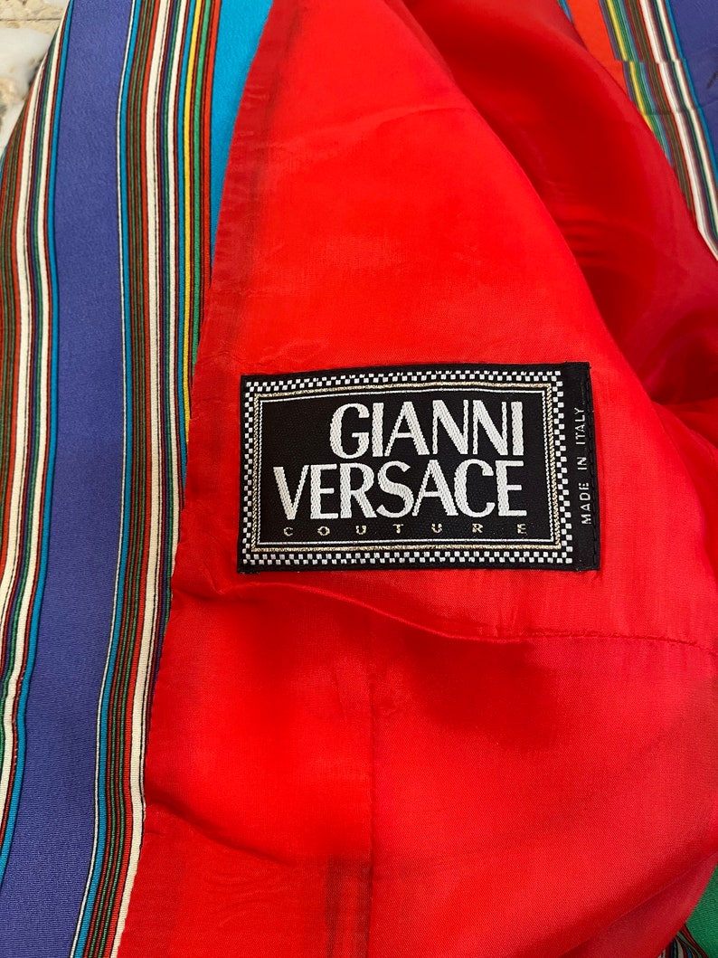 May include: A red fabric with a black label that reads "Gianni Versace Couture" and "Made in Italy". The fabric has a striped pattern in blue, red, yellow, green, and white.