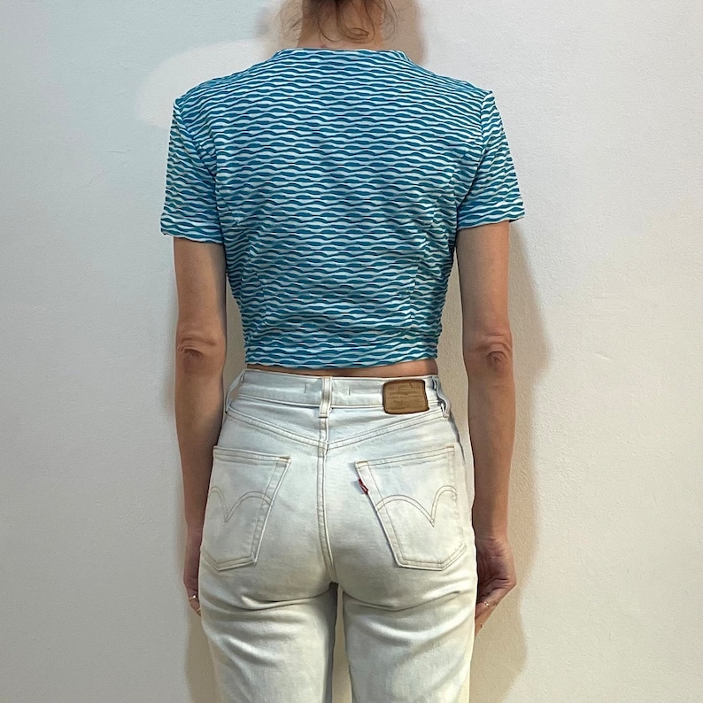 Op de afbeelding: Een blauw en wit gestreept crop top met korte mouwen en een ronde hals. Het topje is gemaakt van een rekbaar materiaal en heeft een golvend patroon.