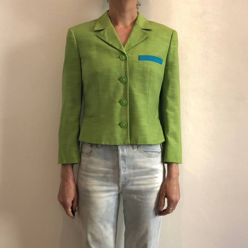 Pode incluir: Um blazer verde curto com uma borda azul no bolso. O blazer tem um fecho de bot&otilde;es e mangas compridas.