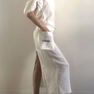 May include: A white linen dress with a scoop neck and a side slit. The dress has a pocket on the left side with a black embroidered design that says "folklore".