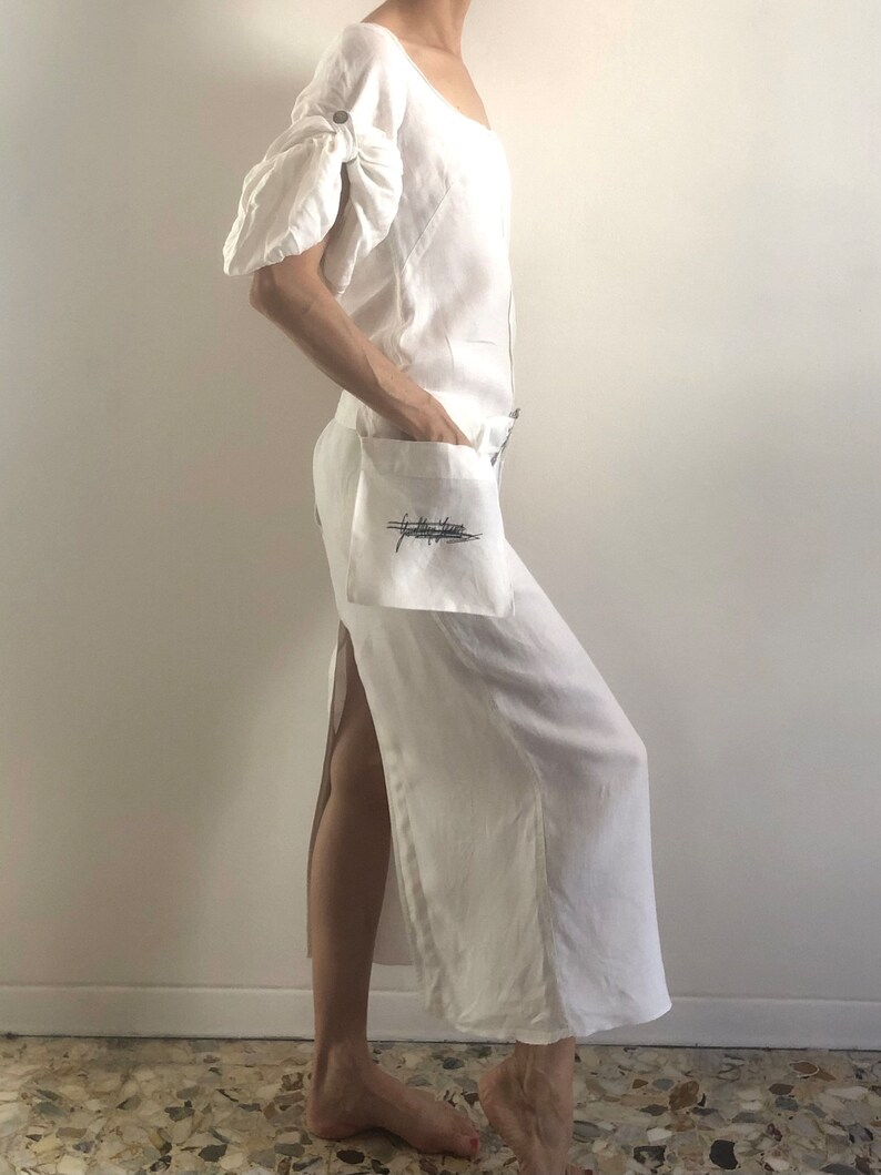 May include: A white linen dress with a scoop neck and a side slit. The dress has a pocket on the left side with a black embroidered design that says "folklore".