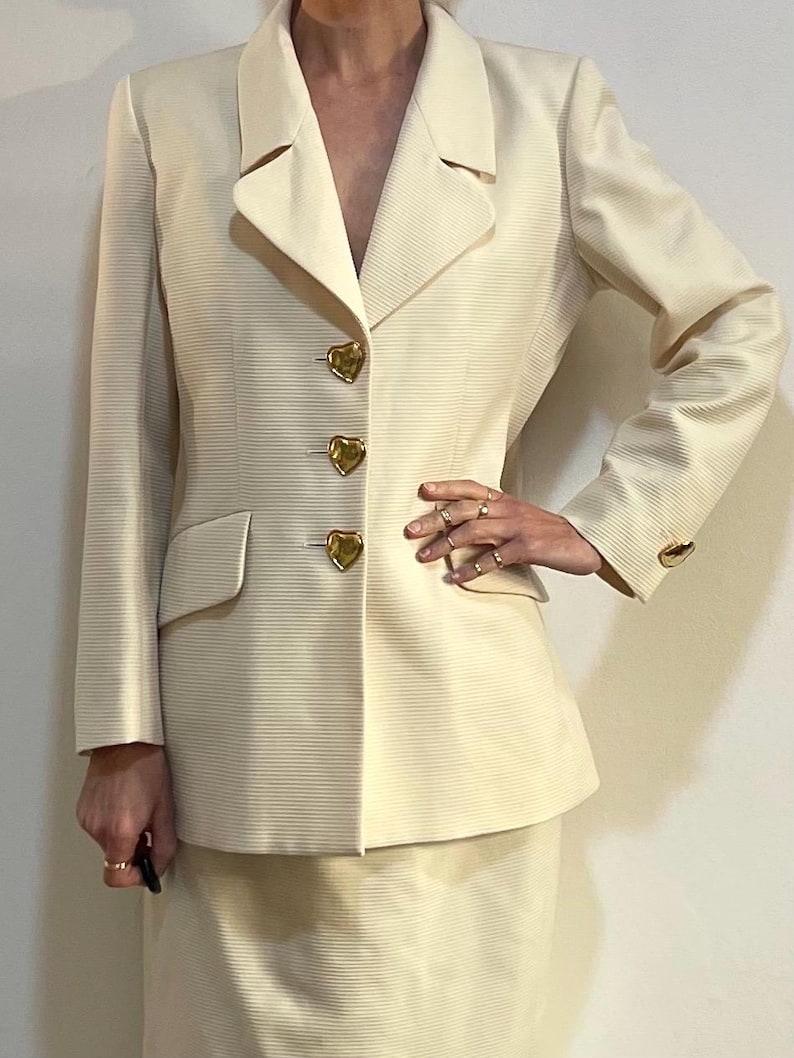 Puede incluir: Traje color crema con blazer y falda. La chaqueta tiene solapas, bolsillos con solapa y botones dorados en forma de coraz&oacute;n. La tela tiene una sutil textura acanalada. El traje es de un estilo cl&aacute;sico y elegante.