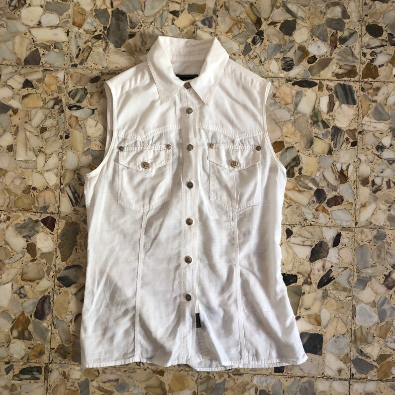 May include: A white sleeveless button-down shirt with a collared neckline and two chest pockets. The shirt has a slightly fitted silhouette and a slightly longer length.