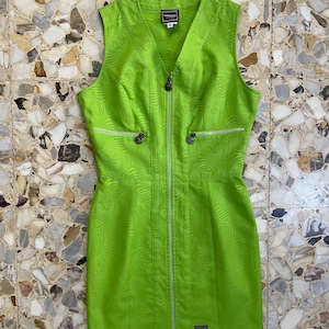 Może przedstawiać: Zielona sukienka bez rękaw&oacute;w z zamkiem błyskawicznym z przodu i wzorem w postaci spirali. Sukienka ma dekolt w kształcie litery V, a na metce widnieje nazwa marki "VERSACE".