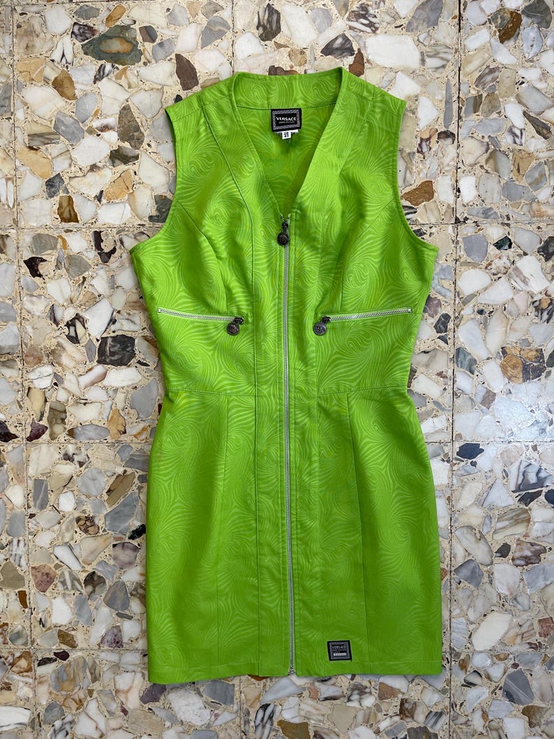 Może przedstawiać: Zielona sukienka bez rękaw&oacute;w z zamkiem błyskawicznym z przodu i wzorem w postaci spirali. Sukienka ma dekolt w kształcie litery V, a na metce widnieje nazwa marki "VERSACE".