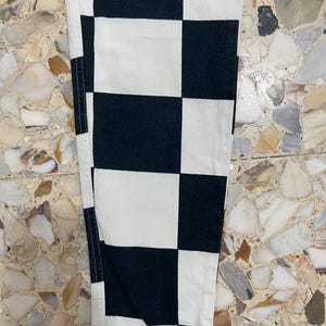 May include: A pair of pants featuring a bold black and white checkered pattern. The garment is constructed with visible vertical seams along the sides and displays a consistent grid of alternating square blocks throughout the fabric surface.
