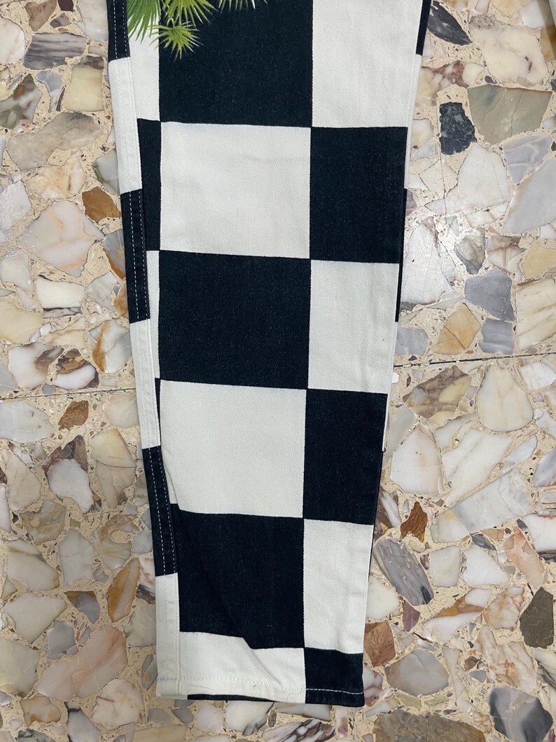 May include: A pair of pants featuring a bold black and white checkered pattern. The garment is constructed with visible vertical seams along the sides and displays a consistent grid of alternating square blocks throughout the fabric surface.