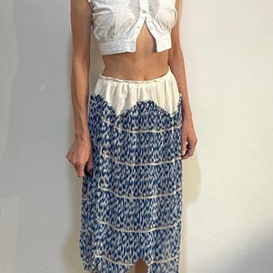 Op de afbeelding: Een witte mouwloze crop top met kanten details en een knoopsluiting. Gecombineerd met een midi-rok met een blauw-wit patroon. De rok heeft een geschulpte zoom, wat zorgt voor een uniek, vintage-ge&iuml;nspireerd ensemble.