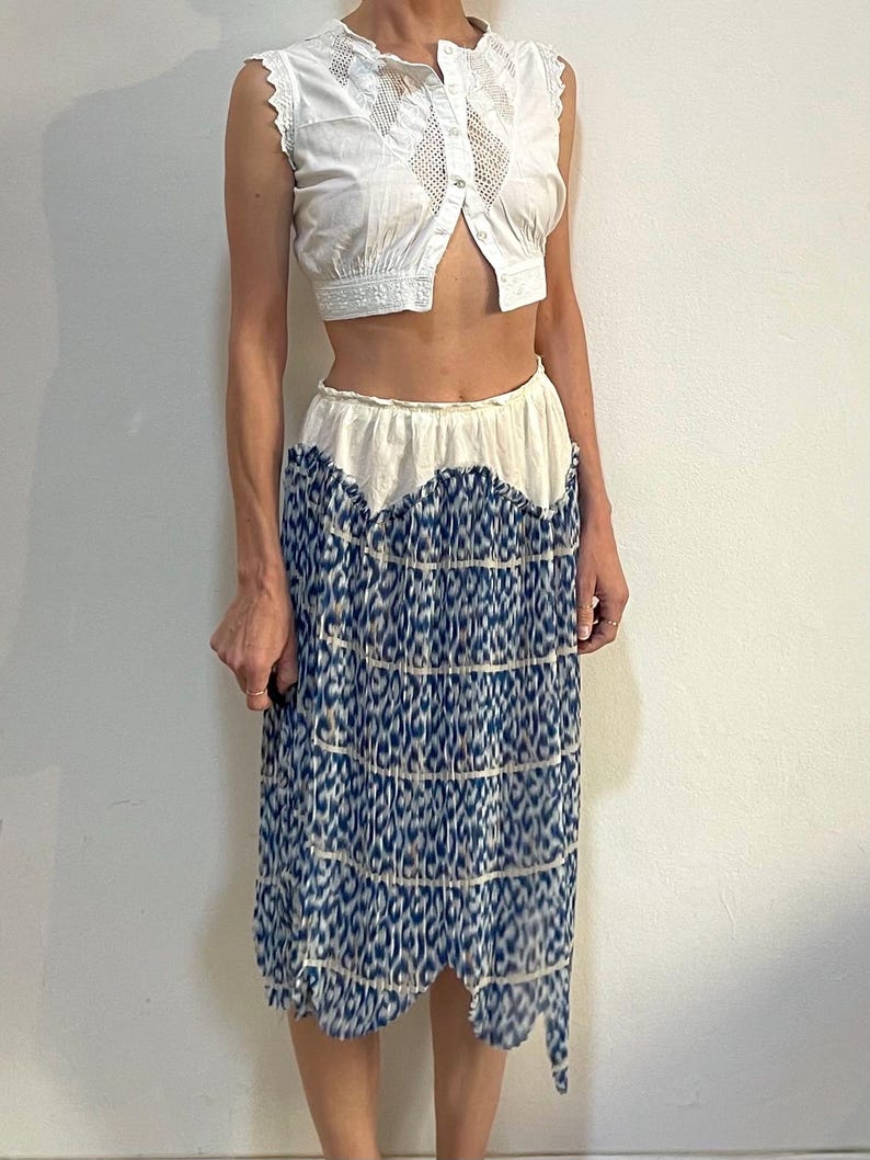 Op de afbeelding: Een witte mouwloze crop top met kanten details en een knoopsluiting. Gecombineerd met een midi-rok met een blauw-wit patroon. De rok heeft een geschulpte zoom, wat zorgt voor een uniek, vintage-ge&iuml;nspireerd ensemble.