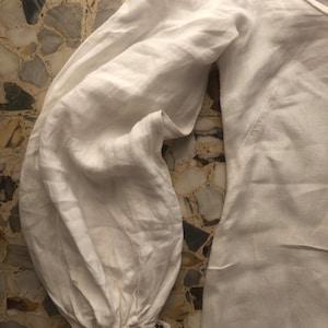 May include: White linen shirt with long sleeves and a gathered cuff detail. The shirt has a relaxed fit and a slightly oversized silhouette.