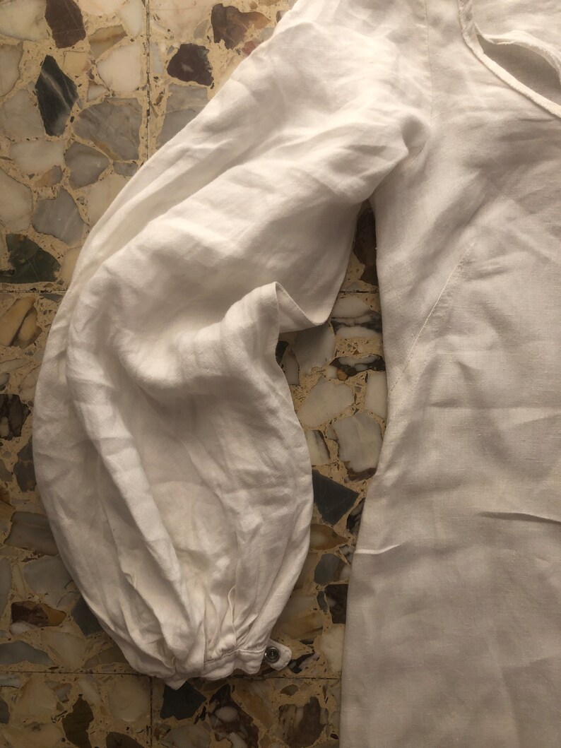 May include: White linen shirt with long sleeves and a gathered cuff detail. The shirt has a relaxed fit and a slightly oversized silhouette.