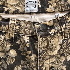 May include: Black denim jeans with a floral print in shades of beige and brown. The jeans have a label that reads "Cavalli Jeans" and "Made in Italy".