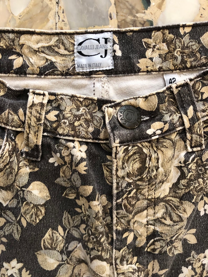 May include: Black denim jeans with a floral print in shades of beige and brown. The jeans have a label that reads "Cavalli Jeans" and "Made in Italy".