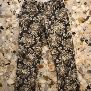 May include: Black denim trousers with a floral print in shades of beige and white. The print features roses and other flowers.