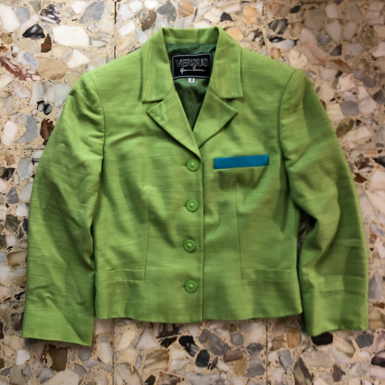 Pode incluir: Um blazer verde com uma borda azul no bolso e uma etiqueta VERSUS Gianni Versace.