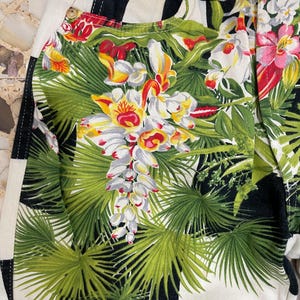 May include: Denim pants featuring a bold black and white checkered pattern overlaid with a vibrant tropical print of green palm leaves and colorful orchids. The garment includes metal rivets and a button closure at the waist with visible stitching.
