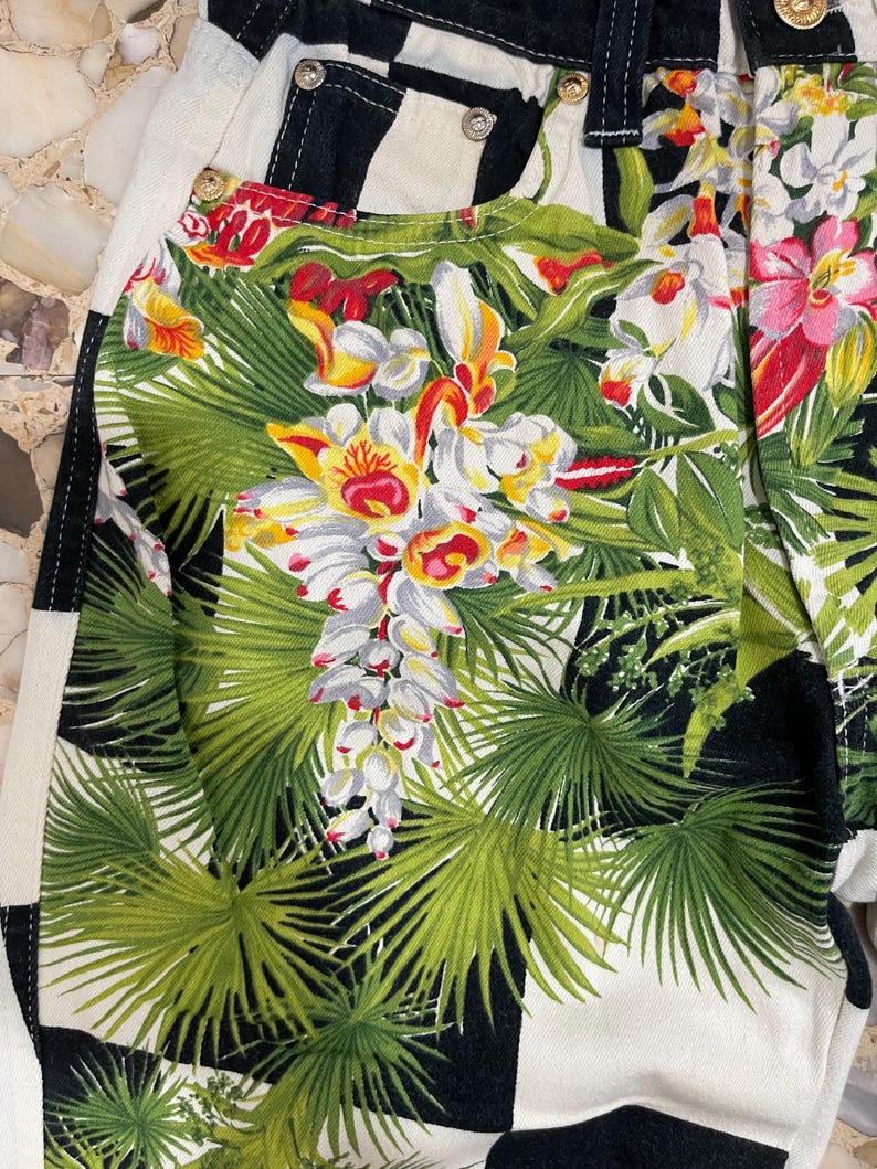 May include: Denim pants featuring a bold black and white checkered pattern overlaid with a vibrant tropical print of green palm leaves and colorful orchids. The garment includes metal rivets and a button closure at the waist with visible stitching.