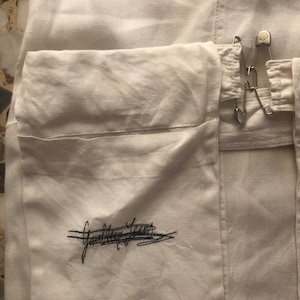 May include: White linen garment with a large pocket on the front. The pocket has a black embroidered signature that reads "Ganni Team". The garment has a safety pin closure.
