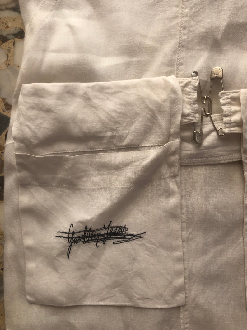 May include: White linen garment with a large pocket on the front. The pocket has a black embroidered signature that reads "Ganni Team". The garment has a safety pin closure.