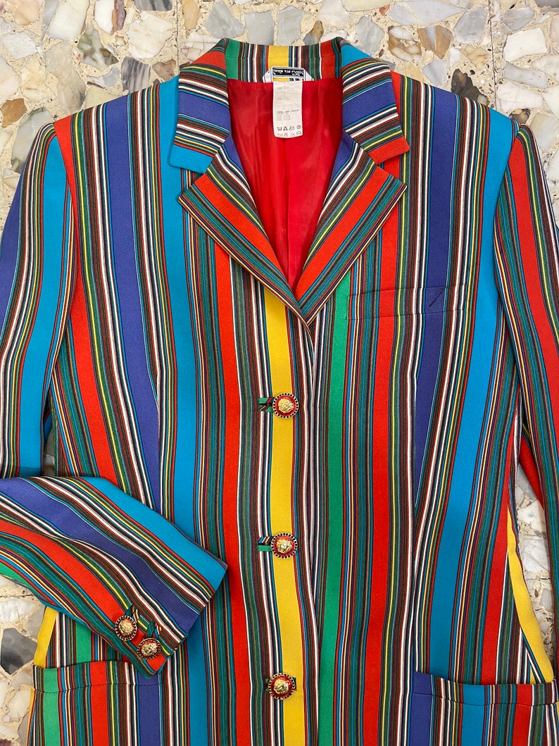 May include: A long-sleeved blazer with a colorful striped pattern in shades of red, blue, yellow, green, and black. The blazer has gold lion head buttons.