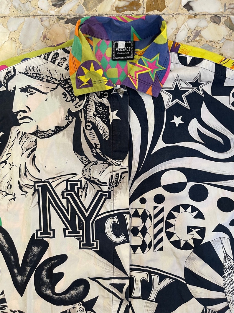K&ouml;nnte beinhalten: Ein wei&szlig;es Hemd mit Kn&ouml;pfen und einem schwarzen, wei&szlig;en und gelben Druck mit der Freiheitsstatue, den Worten "NY" und "Big City" sowie Sternen. Der Kragen ist ein mehrfarbiger Druck mit dem Wort "VERSACE" in einem schwarzen Rechteck.