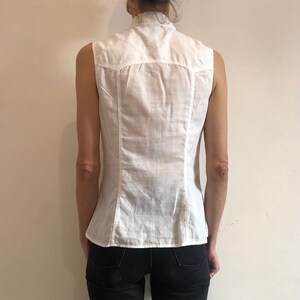 May include: A white sleeveless button-up shirt with a collar. The shirt is made of a lightweight fabric and has a slightly fitted silhouette.