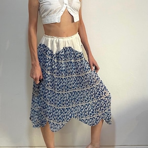 Op de afbeelding: Een witte, mouwloze, cropped top met een geschulpte rand en een knoopsluiting aan de voorkant. Gecombineerd met een vloeiende, knielange rok met een blauw-wit dierenprint ontwerp. De rok heeft een geschulpte zoom.