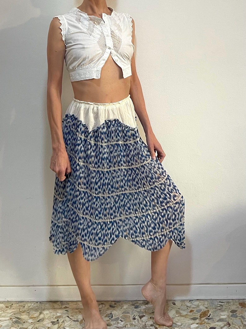 Op de afbeelding: Een witte, mouwloze, cropped top met een geschulpte rand en een knoopsluiting aan de voorkant. Gecombineerd met een vloeiende, knielange rok met een blauw-wit dierenprint ontwerp. De rok heeft een geschulpte zoom.