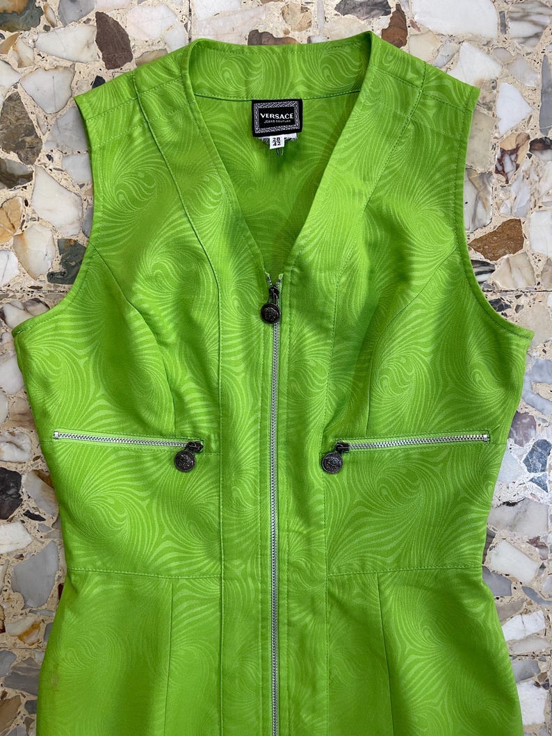 Może przedstawiać: Zielona sukienka bez rękaw&oacute;w z zamkiem błyskawicznym z przodu i wirującym wzorem. Sukienka ma dekolt w kształcie litery V i metkę z napisem "VERSACE" z przodu.