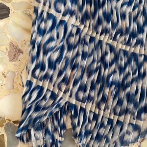 Op de afbeelding: Geplooide stof met een blauw-witte dierenprint. De stof is in verticale lijnen gevouwen, waardoor een textuureffect ontstaat. De print heeft een luipaardachtig patroon, met blauwe vormen op een witte achtergrond.