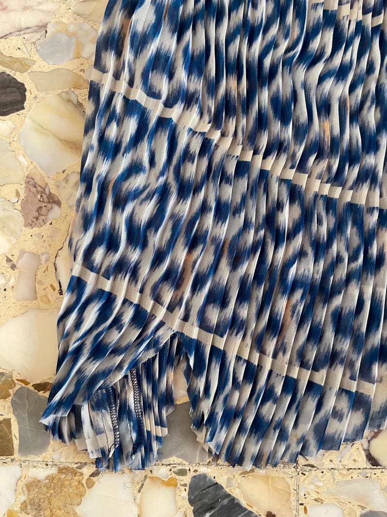 Op de afbeelding: Geplooide stof met een blauw-witte dierenprint. De stof is in verticale lijnen gevouwen, waardoor een textuureffect ontstaat. De print heeft een luipaardachtig patroon, met blauwe vormen op een witte achtergrond.