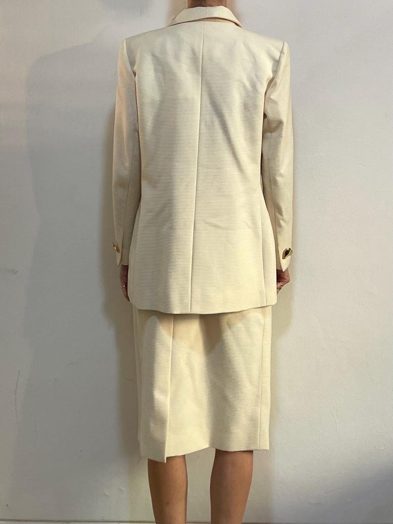 Puede incluir: Traje de falda color crema con tejido acanalado texturizado. El traje incluye una blazer con botones dorados y una falda a juego hasta la rodilla. Un estilo cl&aacute;sico y profesional.