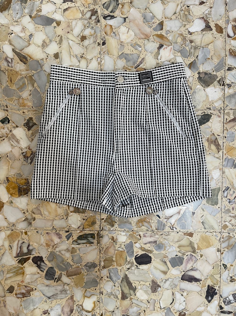 May include: Black and white checkered shorts with decorative silver-toned buttons and zippers. The shorts have a high waist and a small label that reads "VERSACE".