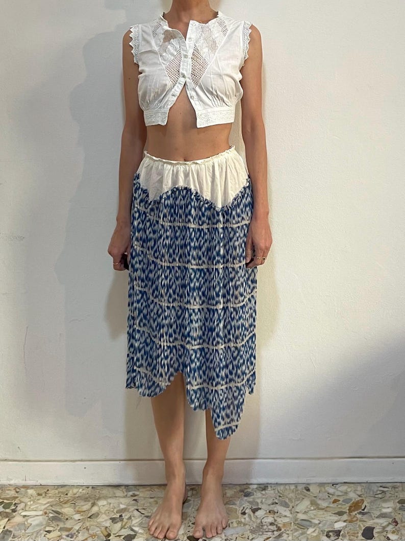 Op de afbeelding: Een witte, mouwloze, knoopsluiting top met kanten details wordt gecombineerd met een blauw-wit patroon rok. De rok heeft een unieke asymmetrische zoom. De outfit is een vintage stijl.