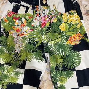 May include: Versace Jeans Couture pants featuring a black and white checkered pattern overlaid with a vibrant tropical floral and palm leaf print. The garment includes a gold-toned button closure, belt loops, and a branded label at the waist.