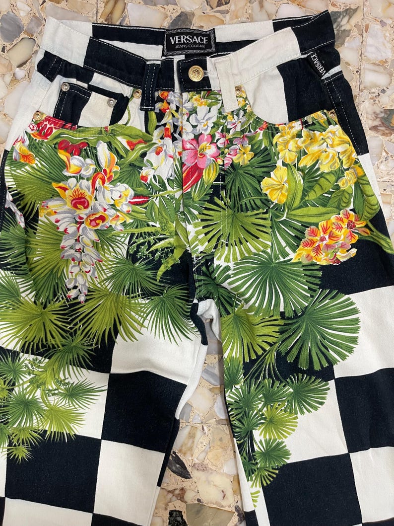 May include: Versace Jeans Couture pants featuring a black and white checkered pattern overlaid with a vibrant tropical floral and palm leaf print. The garment includes a gold-toned button closure, belt loops, and a branded label at the waist.