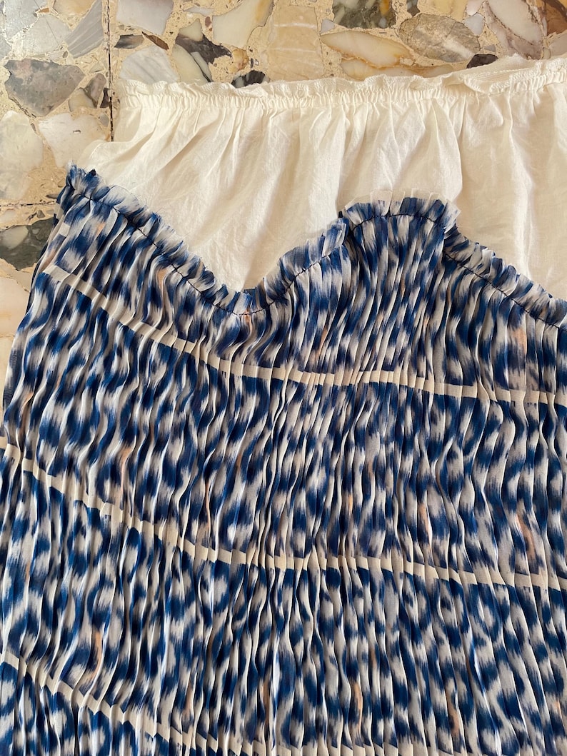 Op de afbeelding: Een blauw-wit patroonstof met een geplooid ontwerp. De stof heeft een getextureerd uiterlijk en een herhalend patroon van blauwe vormen op een witte achtergrond. Een witte, gerimpelde stof is zichtbaar op de achtergrond.