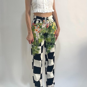 May include: A white sleeveless top with a ruffled hem and button-down front paired with high-waisted jeans featuring a black and white checkerboard pattern overlaid with a vibrant green tropical palm leaf and floral print across the front.