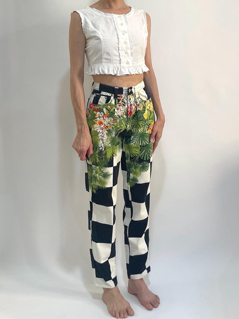 May include: A white sleeveless top with a ruffled hem and button-down front paired with high-waisted jeans featuring a black and white checkerboard pattern overlaid with a vibrant green tropical palm leaf and floral print across the front.