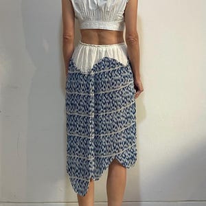 Op de afbeelding: Een witte crop top met kanten rand en een blauw-wit patroon rok. De rok heeft een unieke asymmetrische zoom en een herhalend patroon. Een outfit voor de zomer of lente.