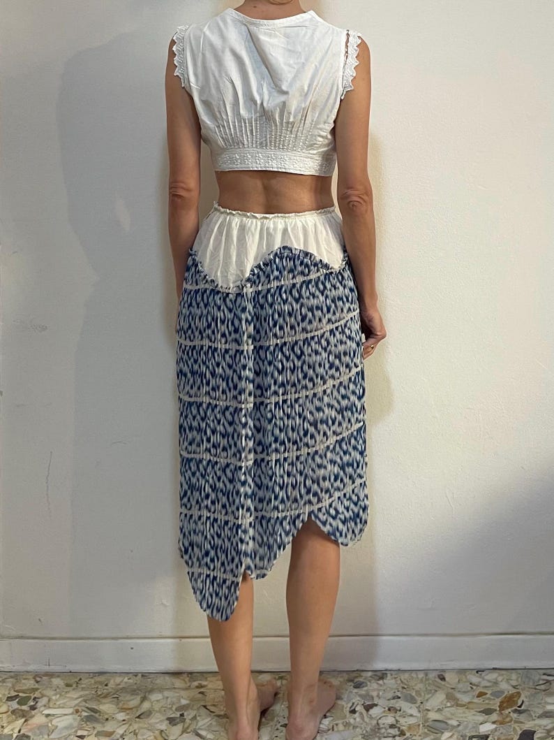 Op de afbeelding: Een witte crop top met kanten rand en een blauw-wit patroon rok. De rok heeft een unieke asymmetrische zoom en een herhalend patroon. Een outfit voor de zomer of lente.