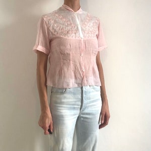 Pu&ograve; includere: Una blusa rosa pallido a maniche corte con colletto bianco e un delicato ricamo rosa intorno allo scollo.