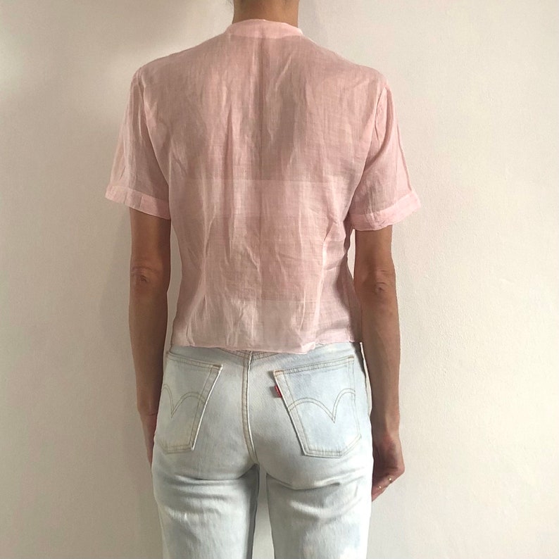 Pu&ograve; includere: Una camicia a maniche corte di colore rosa pallido, trasparente, con bottoni e vestibilit&agrave; rilassata. La camicia &egrave; realizzata in un tessuto leggero e ha un orlo leggermente corto.