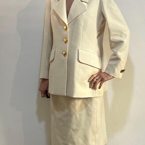 Puede incluir: Traje de falda color crema. La chaqueta tiene solapas con muescas, tres botones dorados en forma de coraz&oacute;n y un bolsillo. La falda llega a la rodilla. El traje tiene una tela acanalada texturizada.